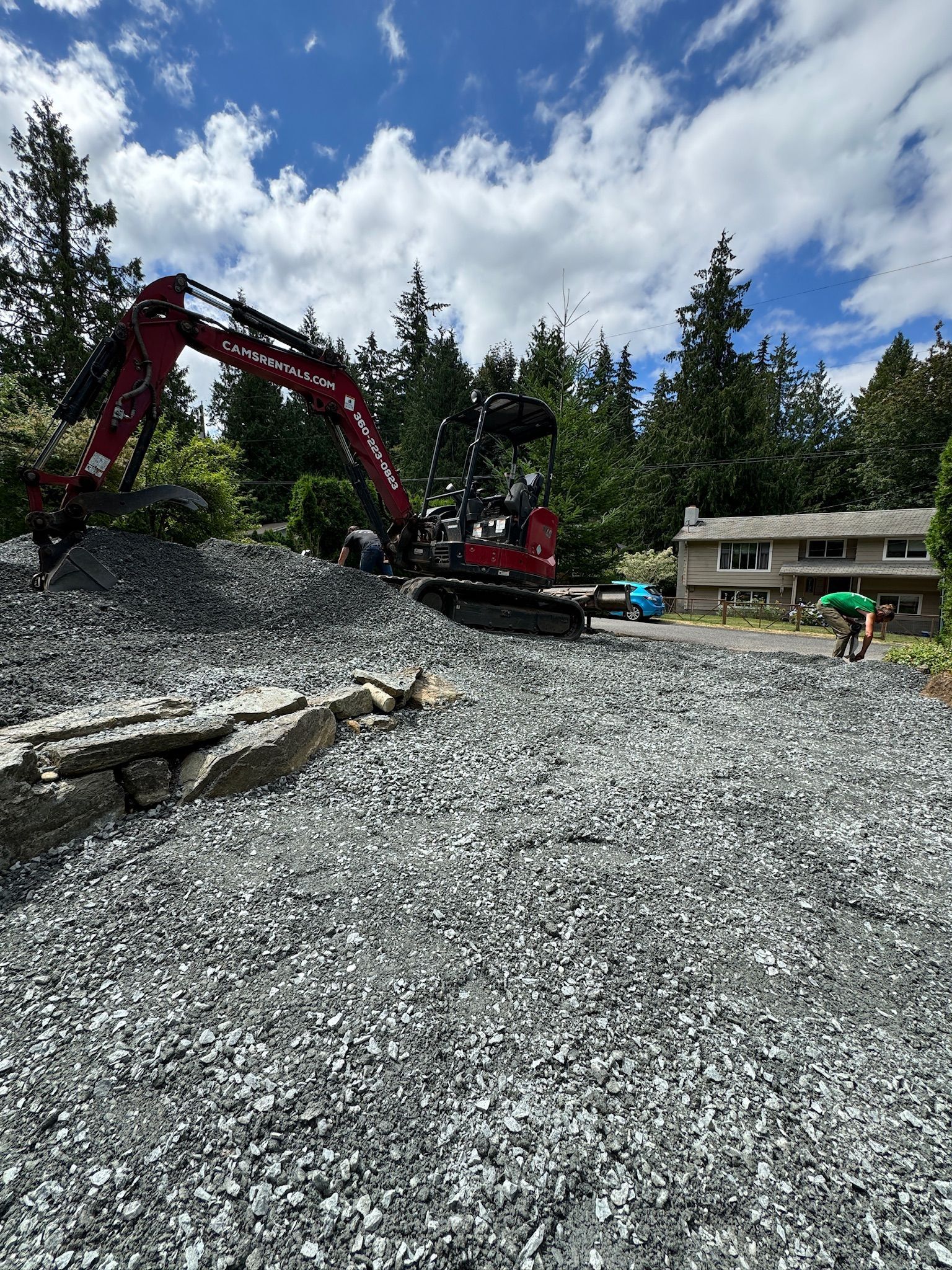 A red excavator is sitting on top of a pile of gravel in front of a house.