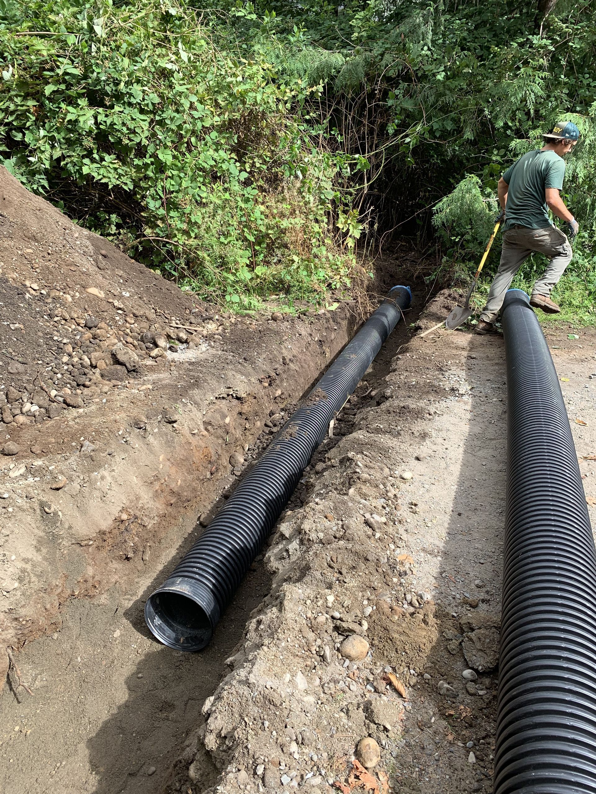 A man is standing next to two black pipes in the dirt.