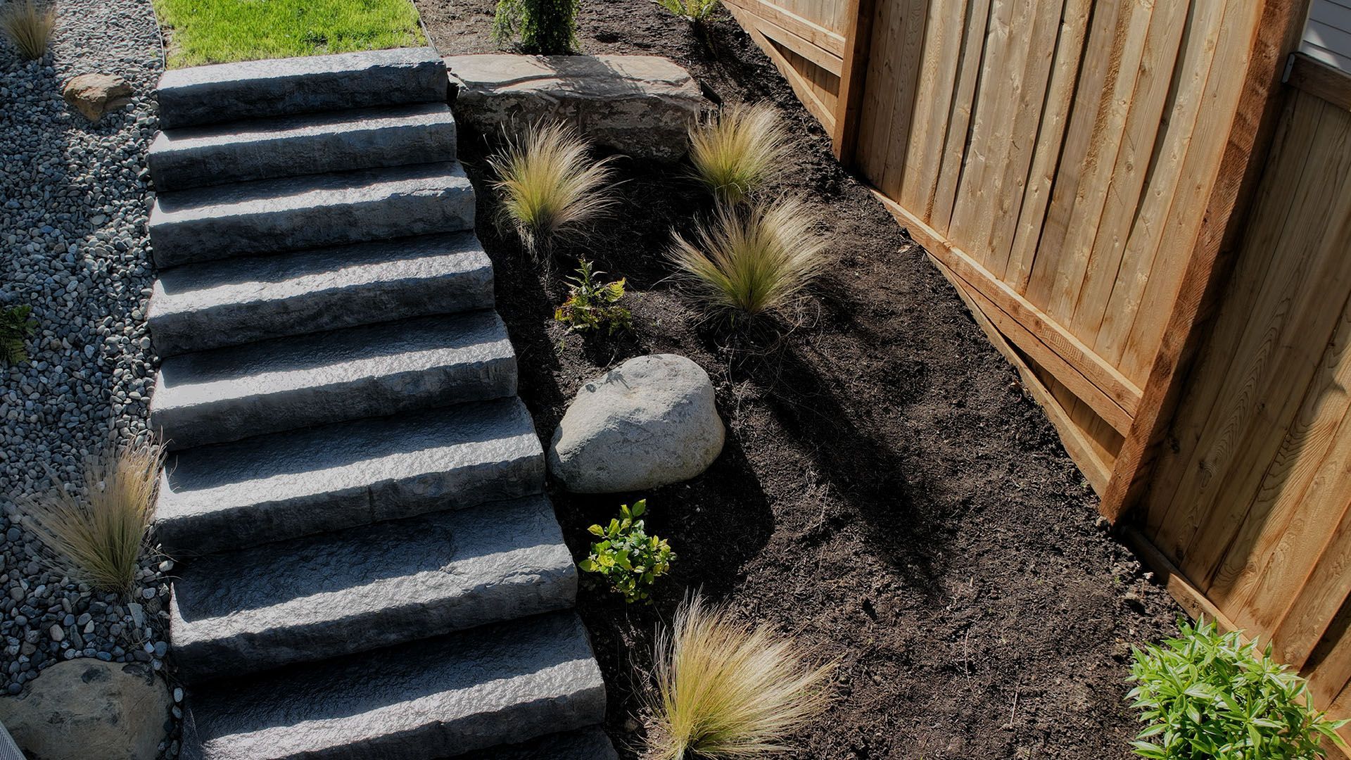 Stone steps lead down to a garden bed with brown mulch, ornamental grass, and a large boulder next to a wooden fence.