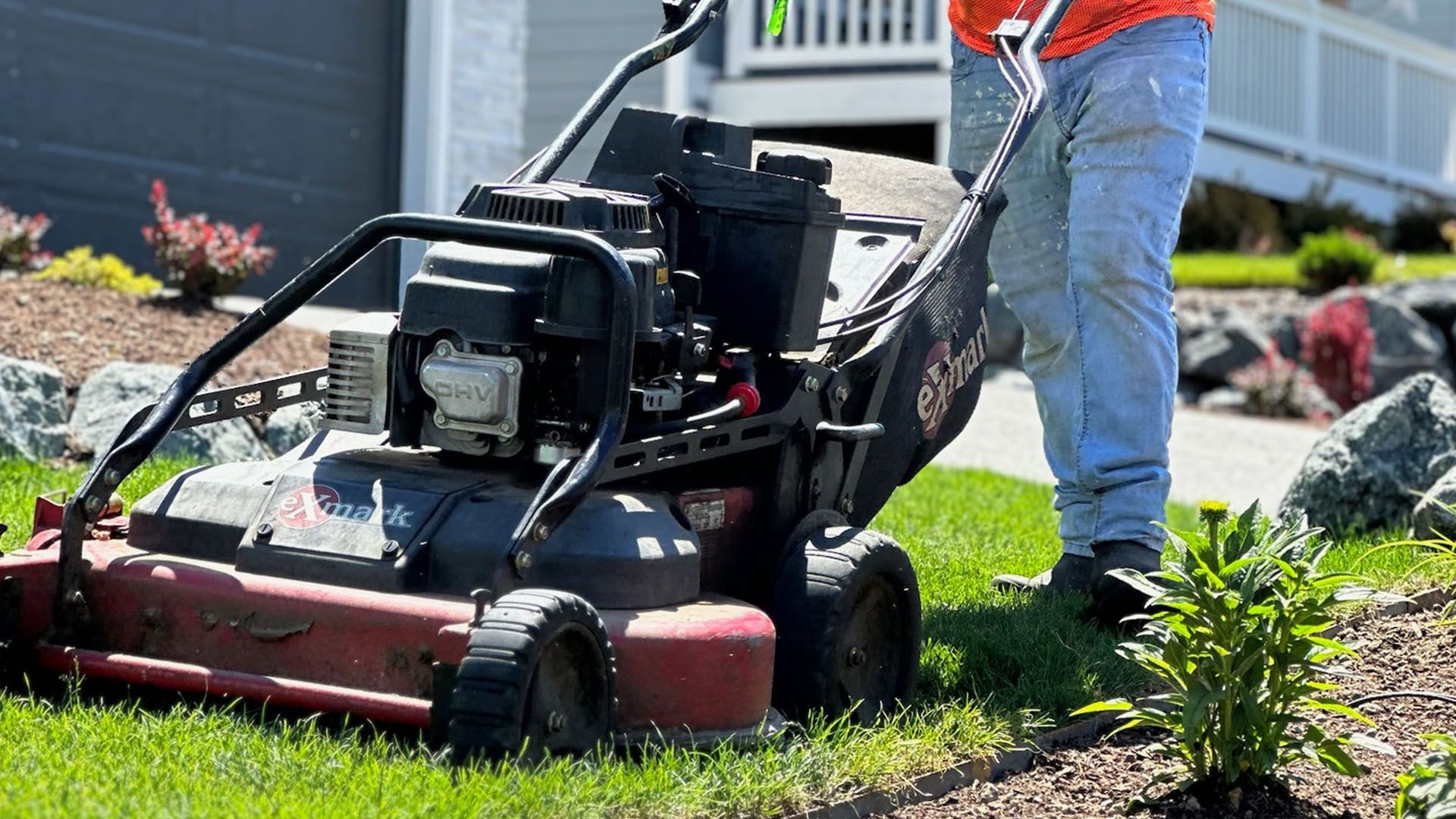 Person mowing a lawn with a black and red lawnmower in a suburban yard.