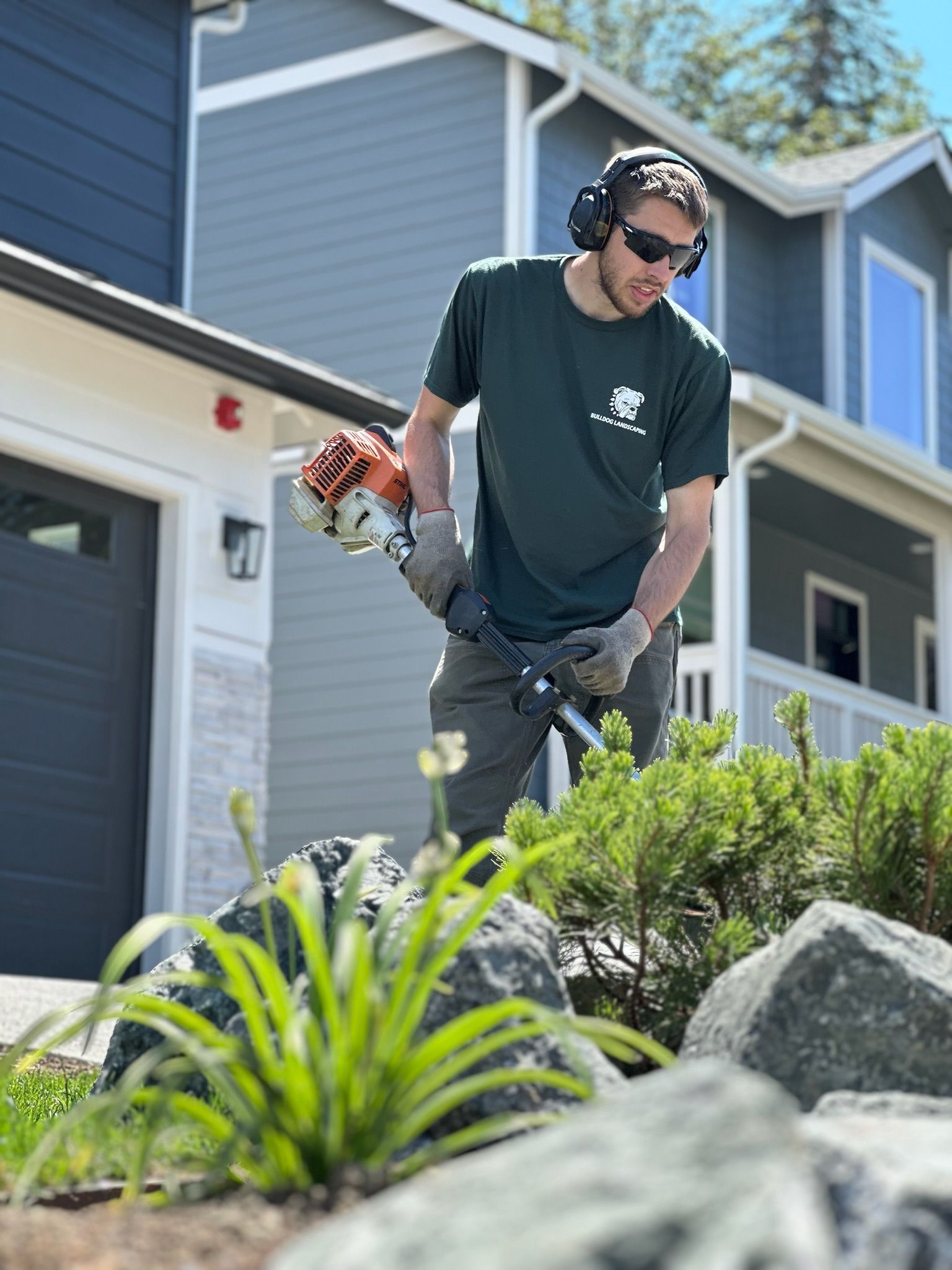 A man wearing headphones and sunglasses is cutting grass in front of a house