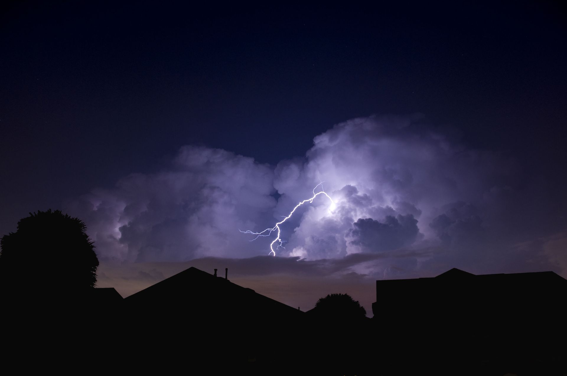 Lightning flashes through dark clouds over silhouetted rooftops at night.