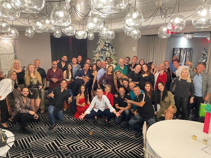 Group of people in a festive room with a decorated tree. Some are smiling, posing. Overhead lights and a round table.