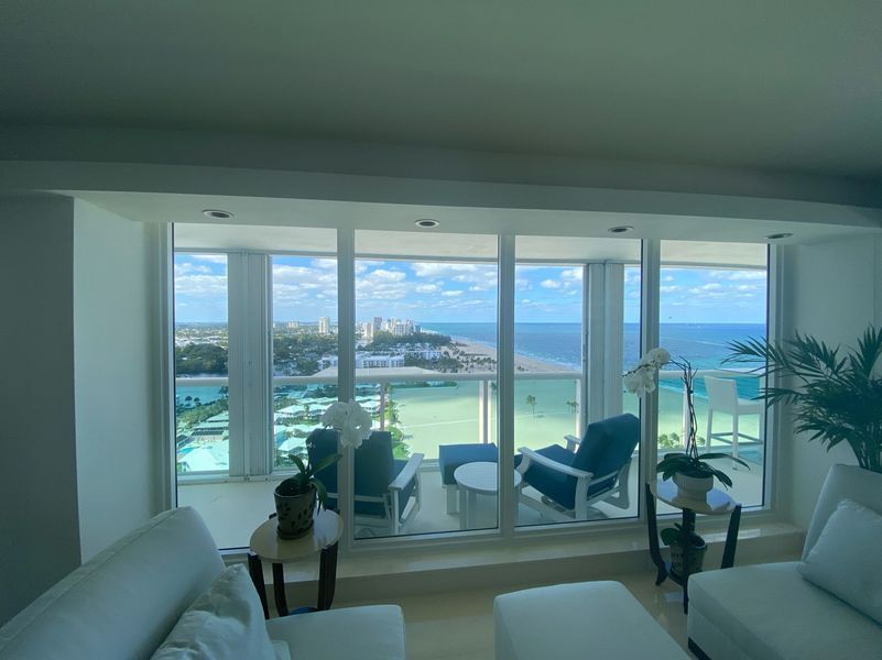 Bright living room with ocean view, white furniture, blue chairs, and potted plants by large windows.