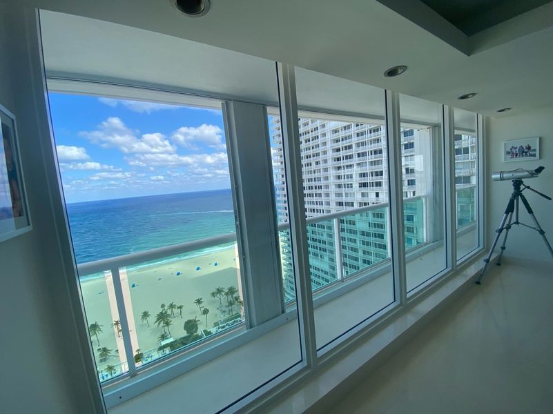 Ocean view from tall windows. Blue sea, sky, and beach. Telescope on a tripod.