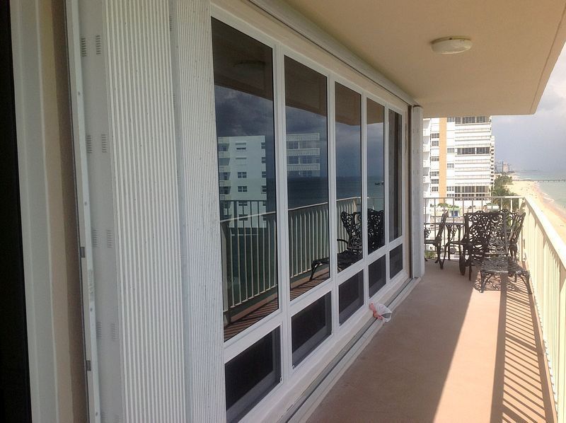 Balcony with floor-to-ceiling windows overlooking the ocean. White shutters and patio furniture are visible.