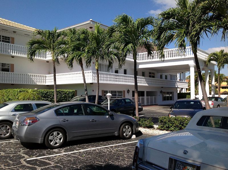 White building with balconies, palm trees, and parked cars in a parking lot on a sunny day.