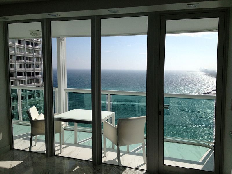 Balcony overlooking ocean; white table, chairs, glass doors, sunny day, building.