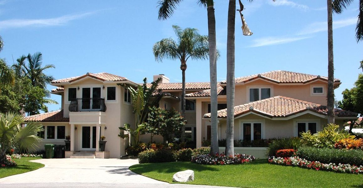 Large beige house with tile roof, palm trees, and well-manicured lawn under a blue sky.