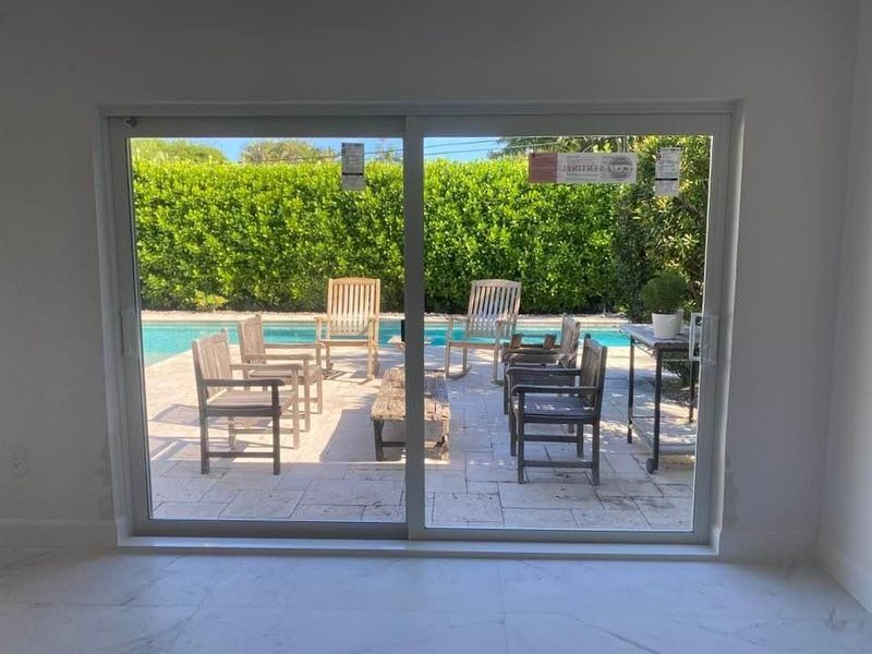 Sliding glass door frames a patio with chairs around a pool, green hedge in the background.