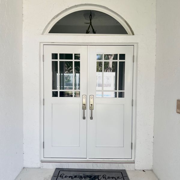 White double doors with glass panels and arched transom, set in a white wall with a welcome mat.