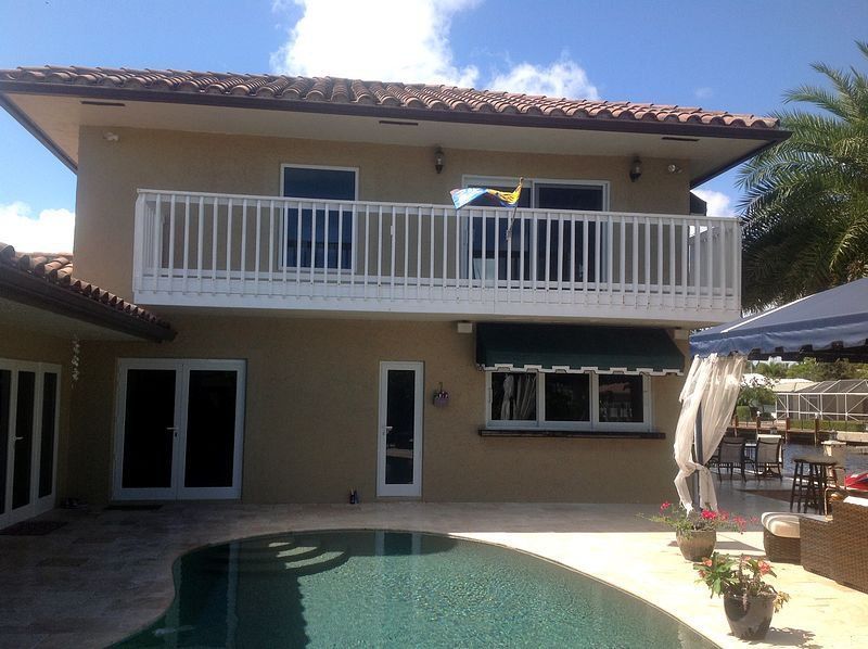 Two-story house with balcony overlooking a pool, tan stucco exterior, white railing, blue sky.