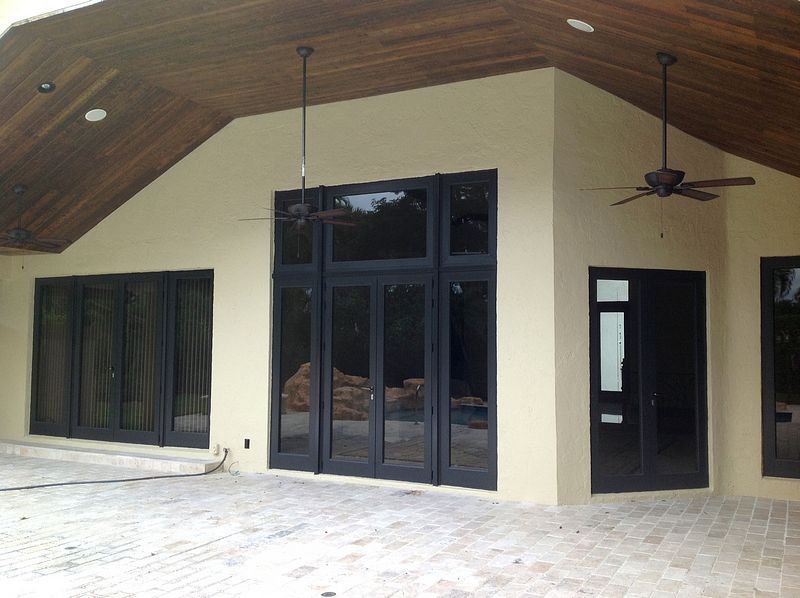 Exterior view of a beige building with black framed windows and doors, wood ceiling, and a brick patio.