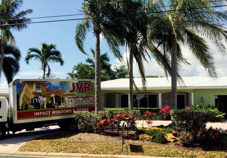 JVR truck parked in front of a light green house with palm trees, impact windows advertised on the truck.