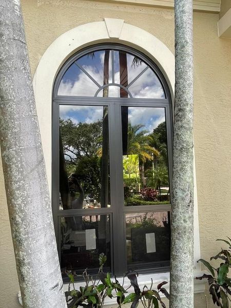 Arched window with dark frames set in a light-colored building, surrounded by trees reflecting outside.