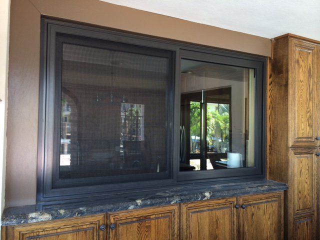 Dark-framed sliding window with screen over a countertop, above wooden cabinets, with a brown trim and wall.