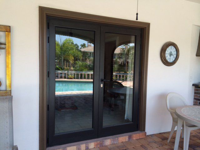 Black double doors with glass panels reflecting a pool, brown frame, on a brick patio.