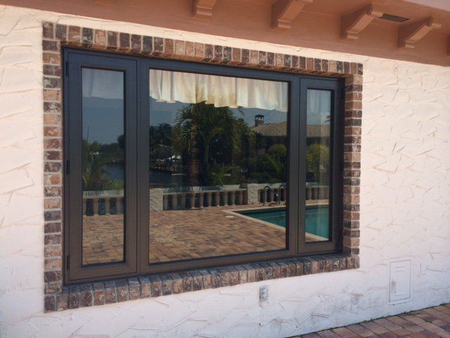 Dark-framed window in brick surround reflects a pool, patio, and palm trees.