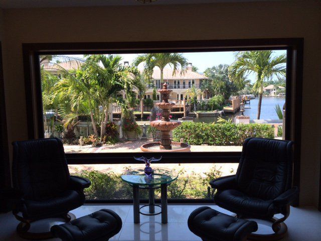 Two black recliners face a large window with a view of a fountain and water canal in a tropical setting.