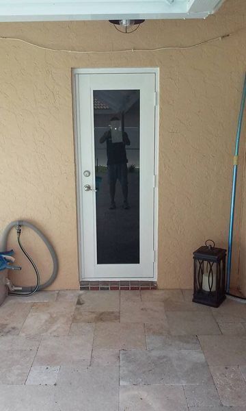 White glass door set in tan stucco wall on a stone patio. A person is reflected in the door.