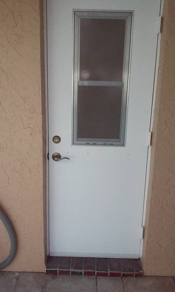 White door with window, set in beige wall. Door has handle and lock. Ground is brick and tile.