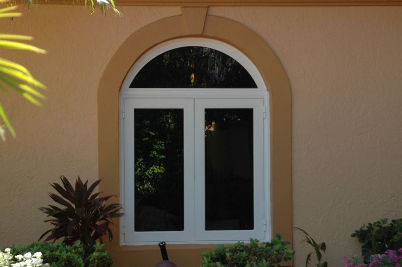 White arched-top window framed in beige, set in tan stucco wall with landscaping.