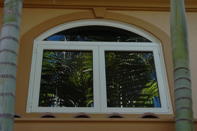 White arched window reflecting lush green foliage, set in tan wall.