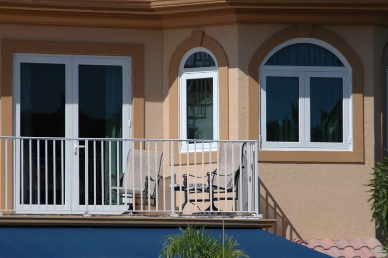 Balcony with white railing, doors, and windows, chairs. Tan building, blue awning.