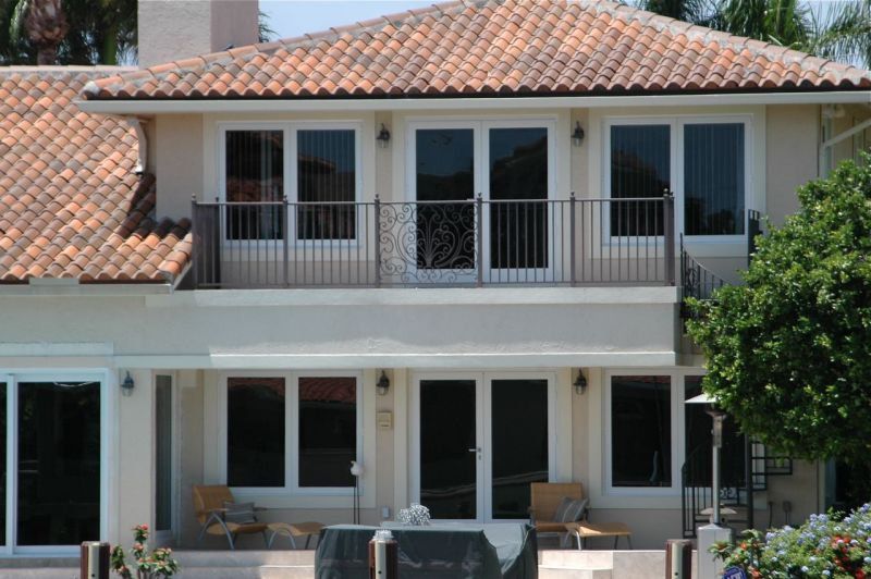 Two-story house with tiled roof, balcony, and large windows.  Cream walls, brown railing, and lush greenery.