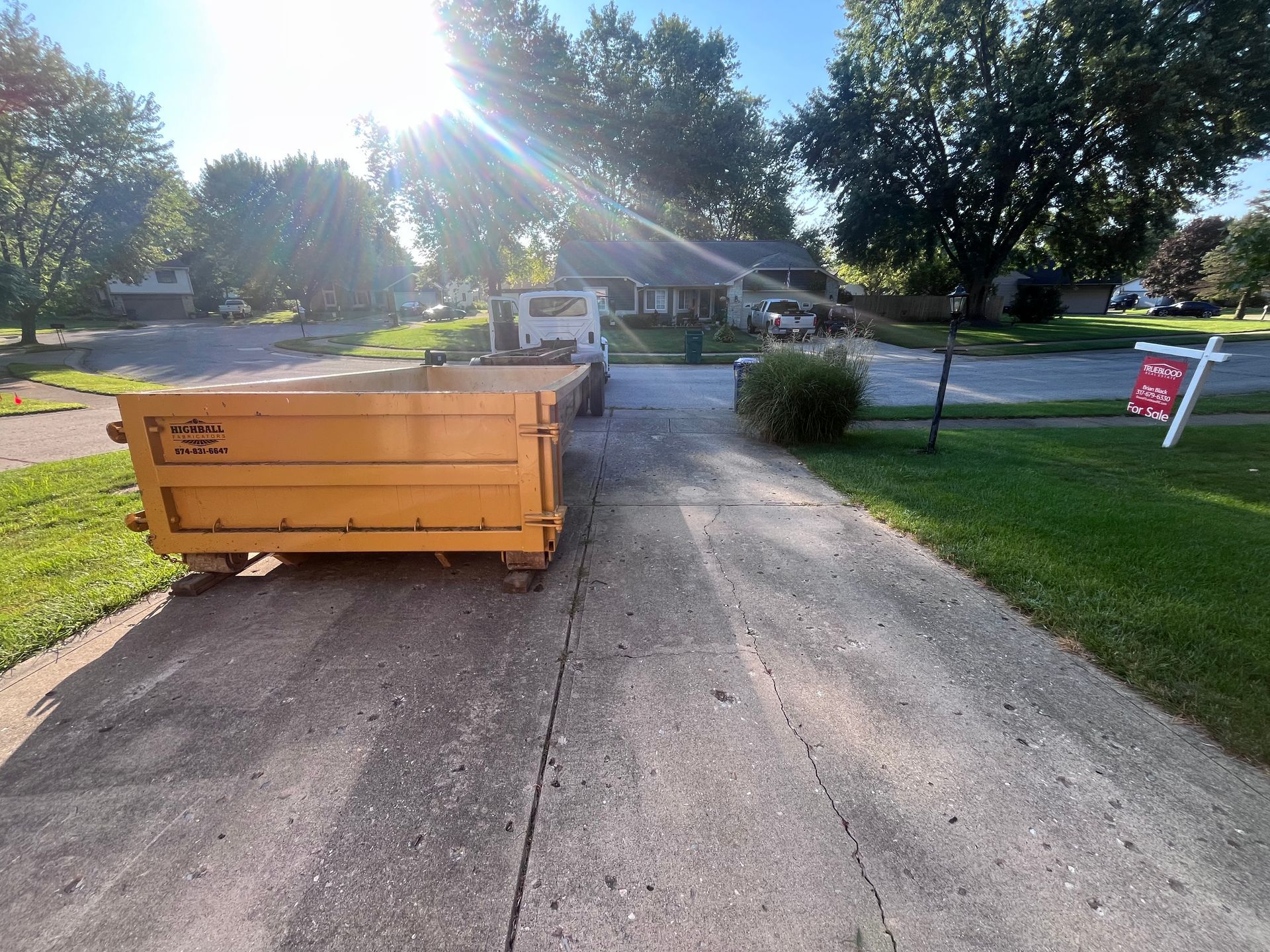 Yellow dumpster sits in a driveway, with a house in the background. Sunlight shines brightly.