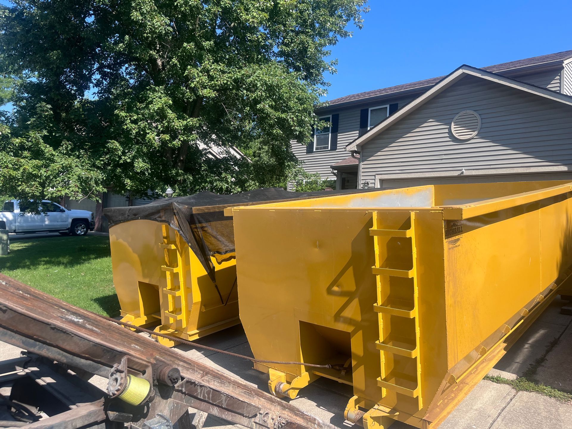 Two yellow dumpsters sit on a driveway in front of a house on a sunny day.
