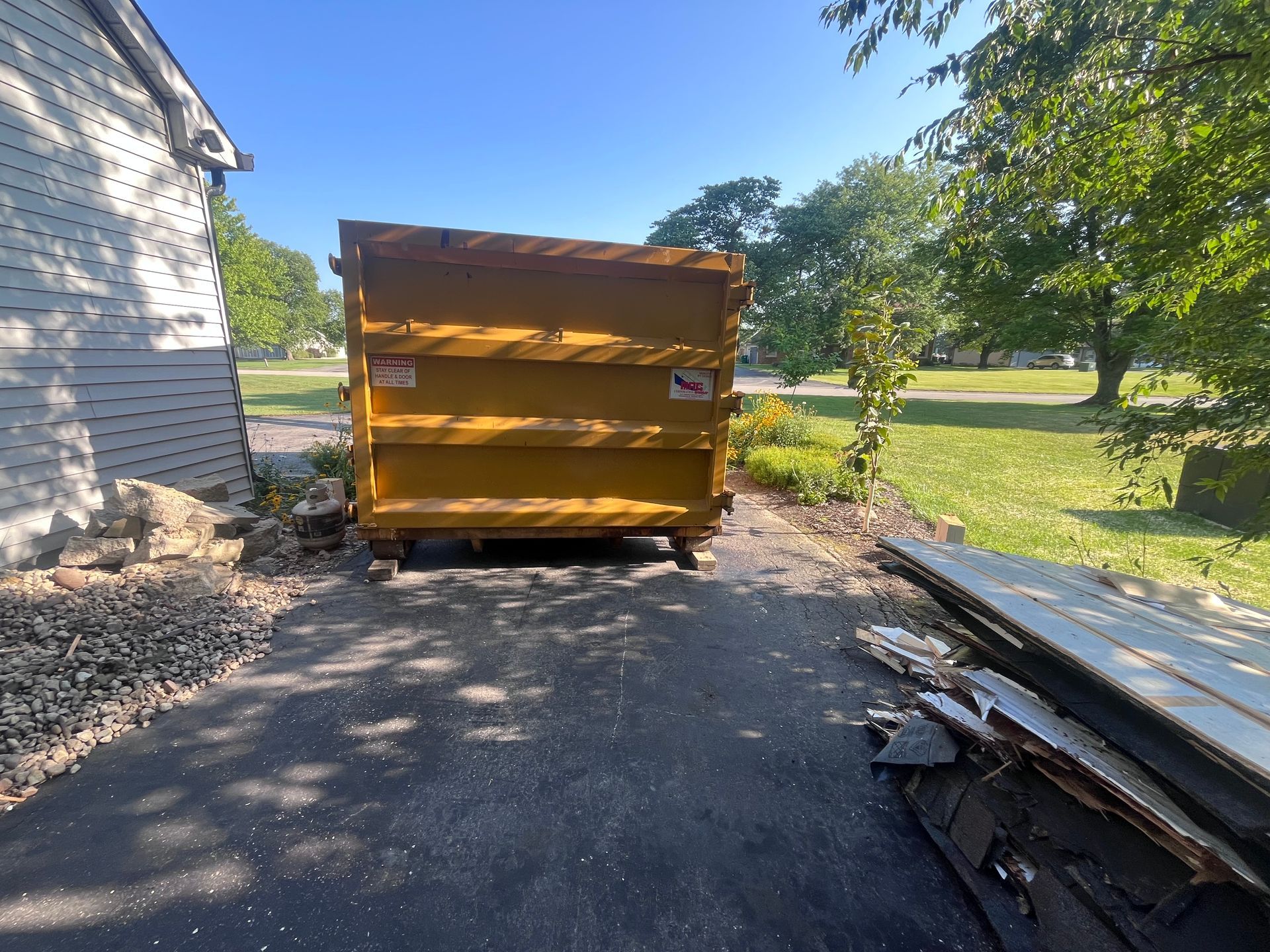 Yellow dumpster on asphalt driveway next to a house with debris and trees in the background.