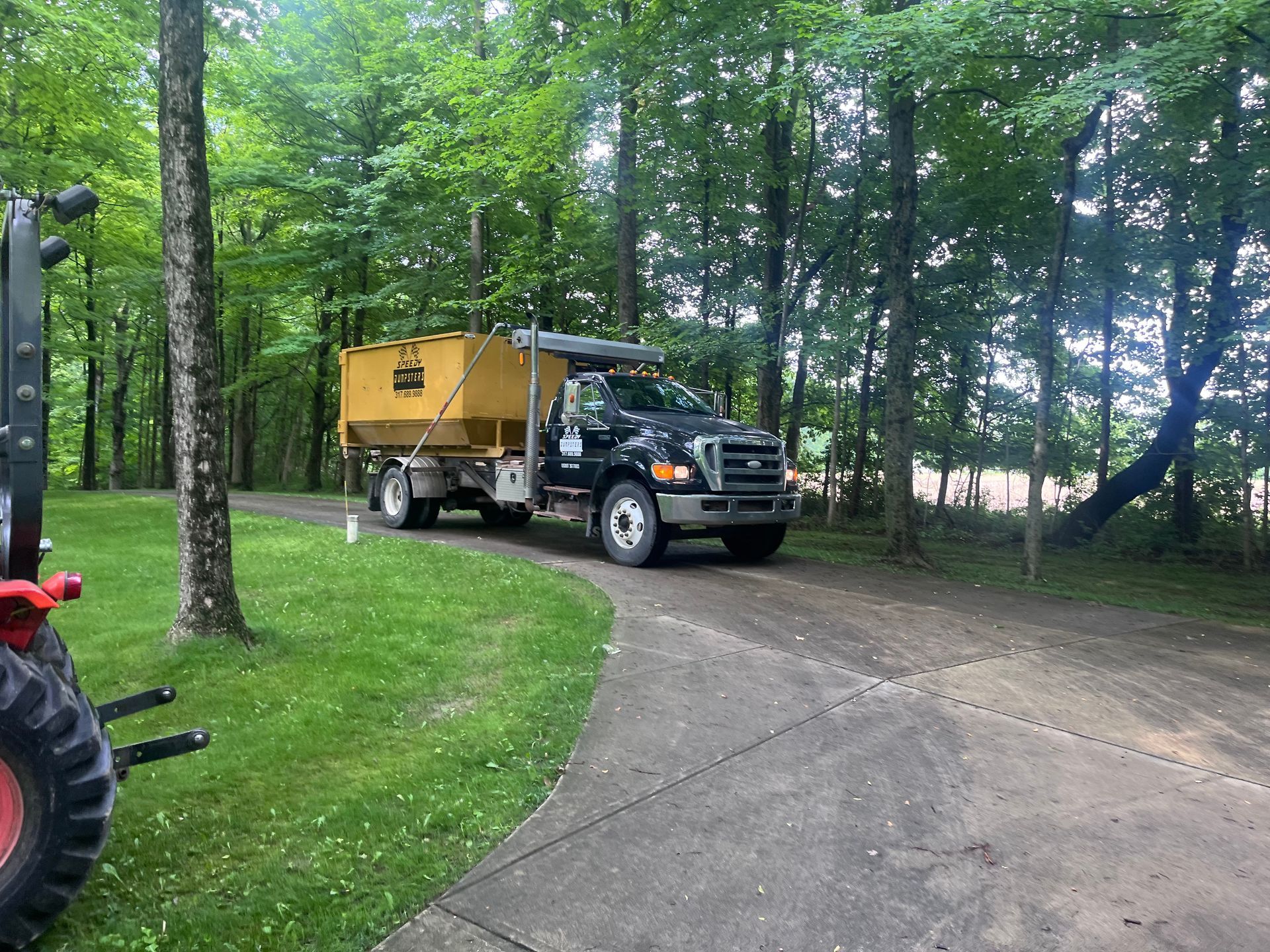 Yellow dump truck on a driveway, next to a red forklift, in a wooded area.