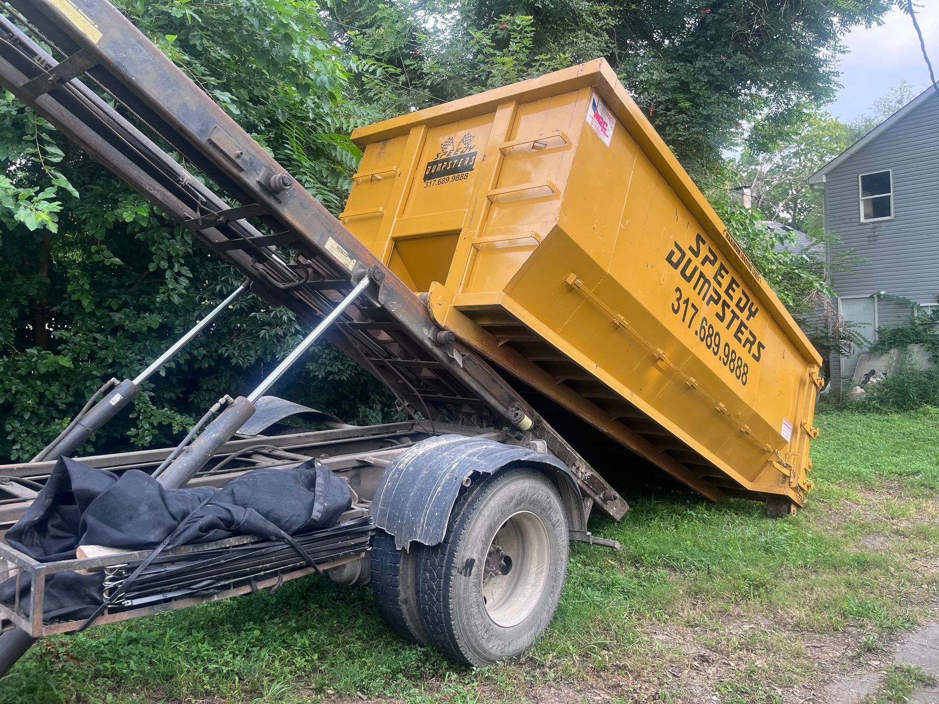 A yellow dumpster being tilted by a tow truck on a grassy area; 