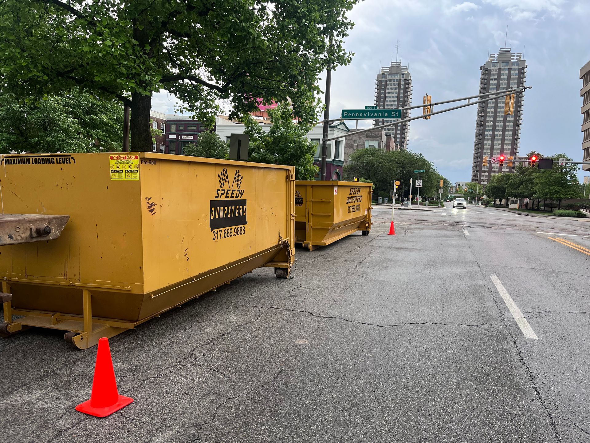 Two yellow dumpsters block a wet city street, orange traffic cone in foreground, tall buildings in background.