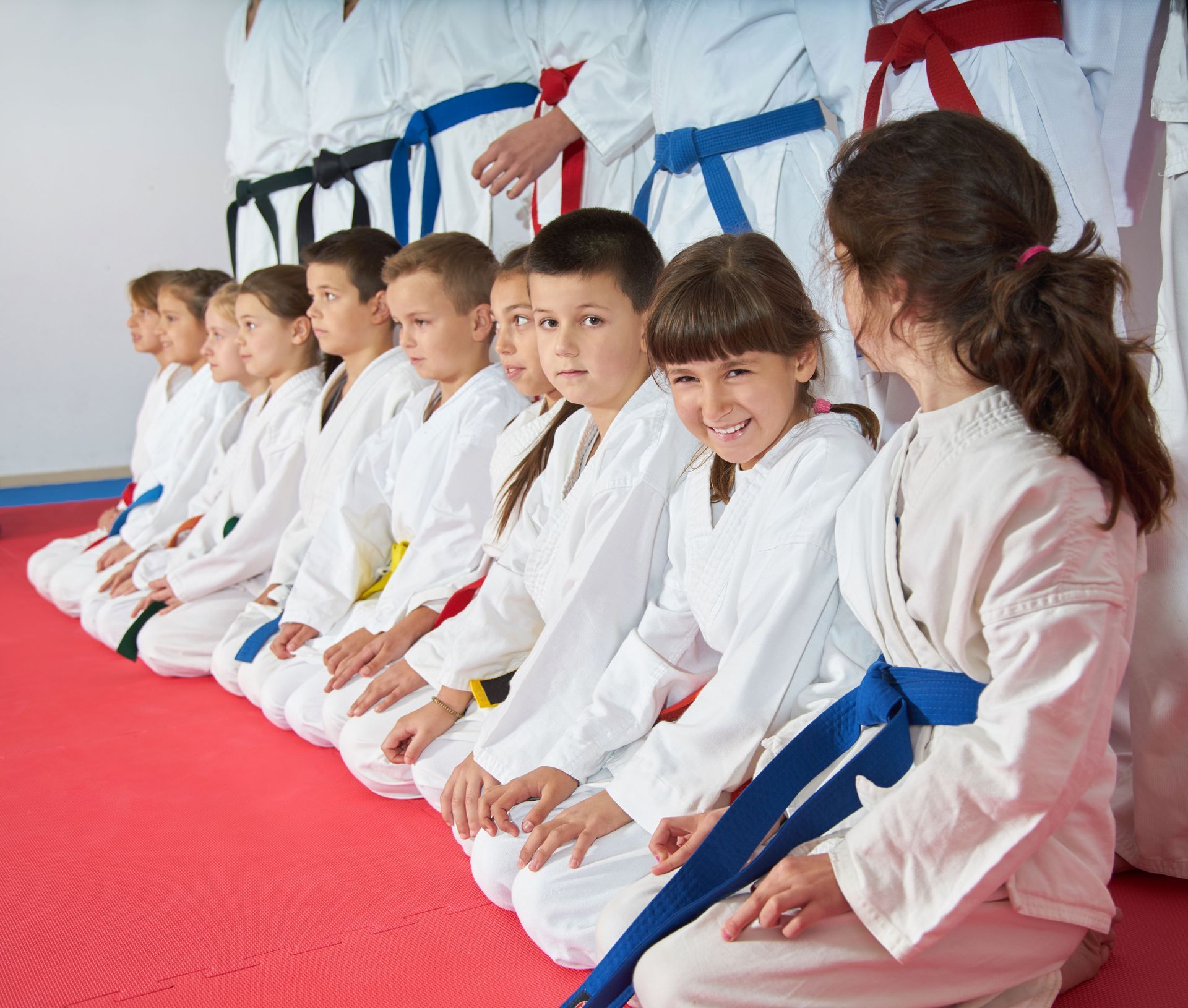 Children in karate uniforms kneeling on red mats, various colored belts, instructor behind them.