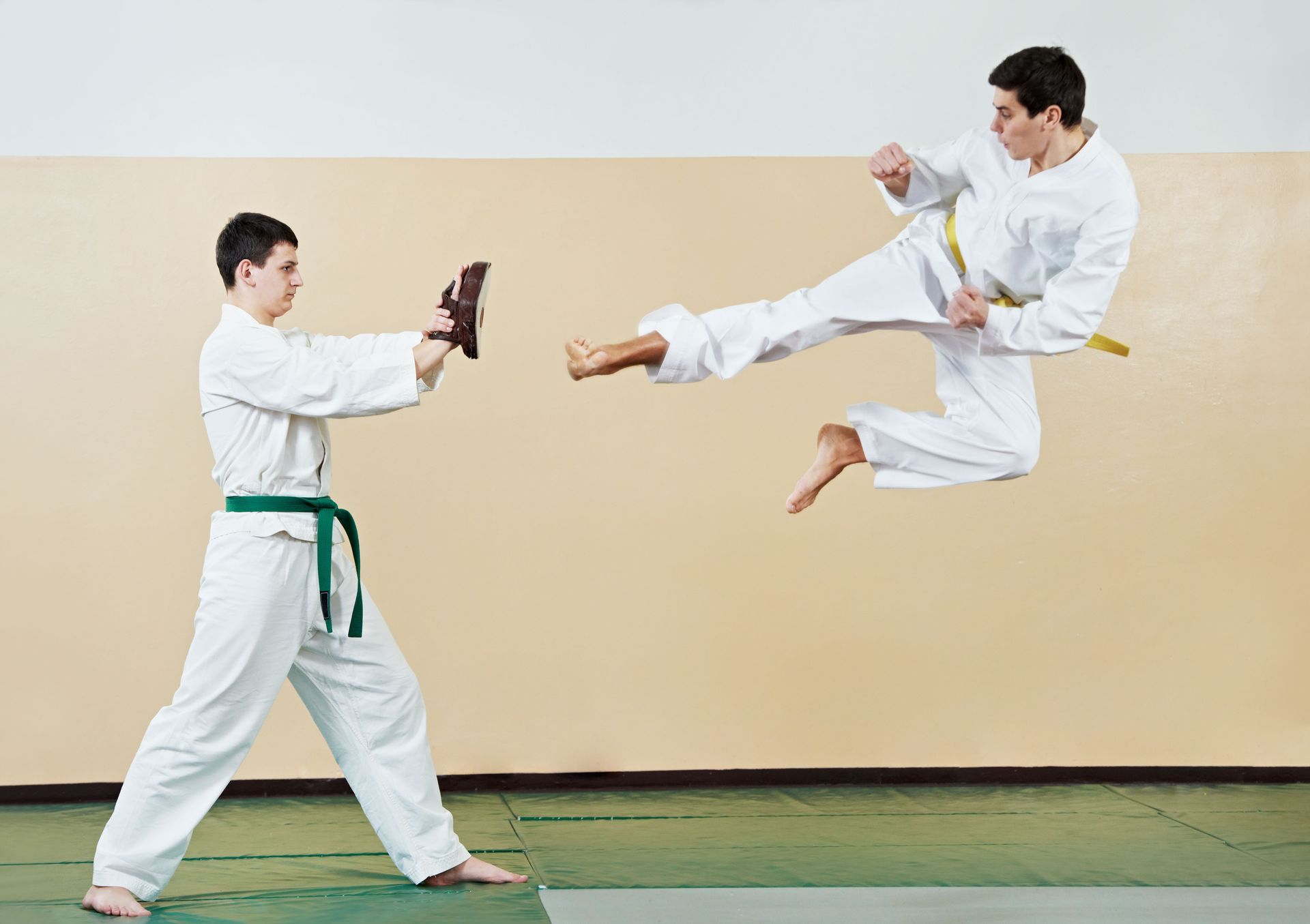 Two people in karate uniforms practicing a flying kick and board break, indoors.