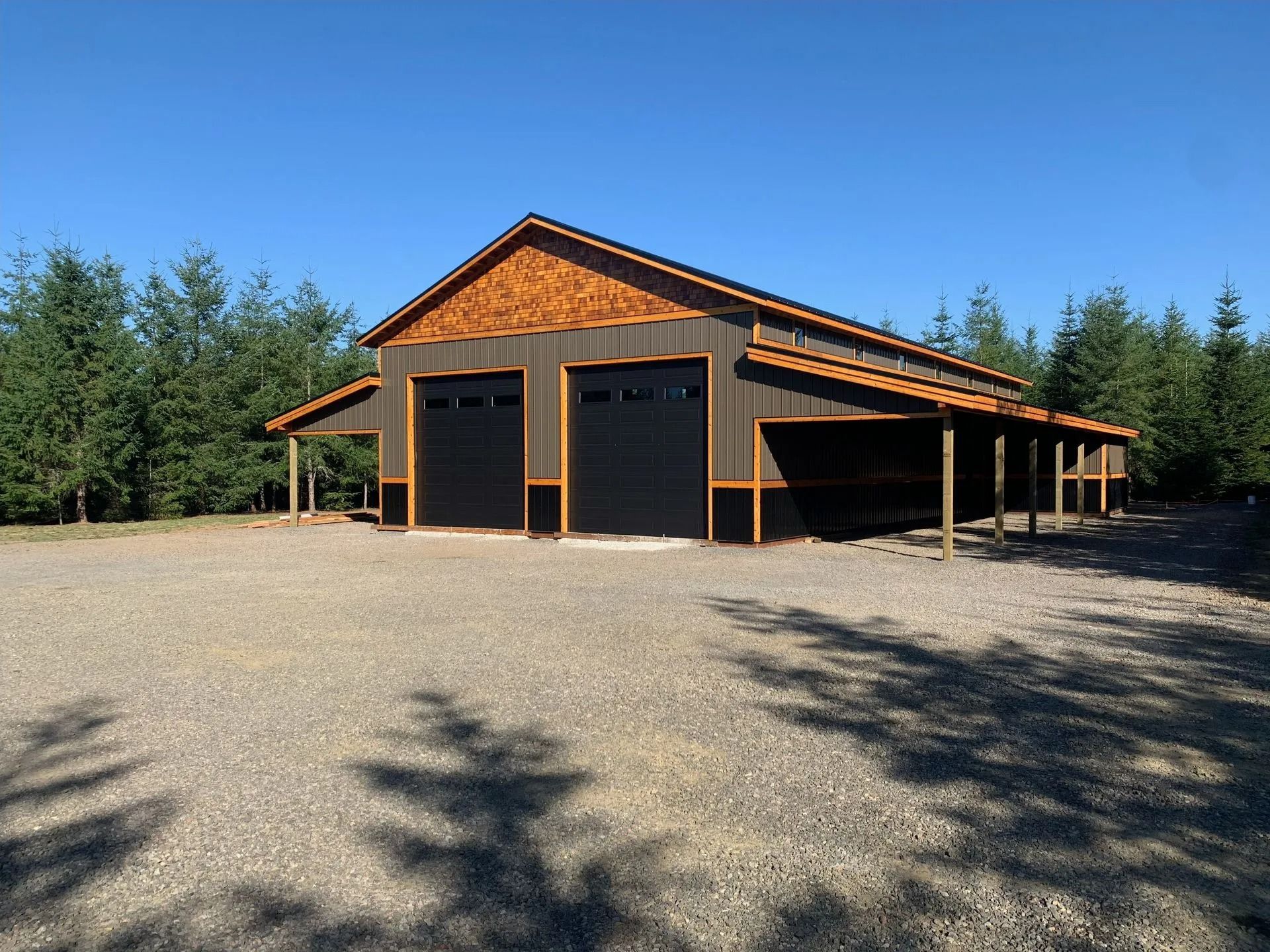 Two-bay, dark-sided barn with black doors and a wood-paneled roof, set on gravel with trees in background.