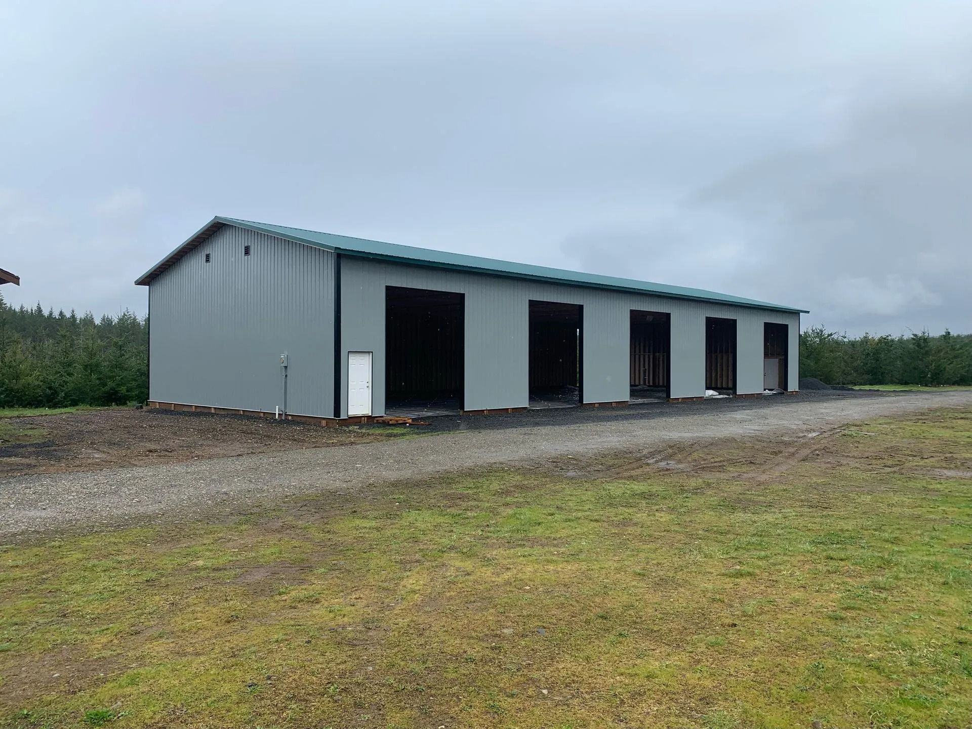 Gray metal building with multiple garage door openings on a gravel driveway, cloudy sky.
