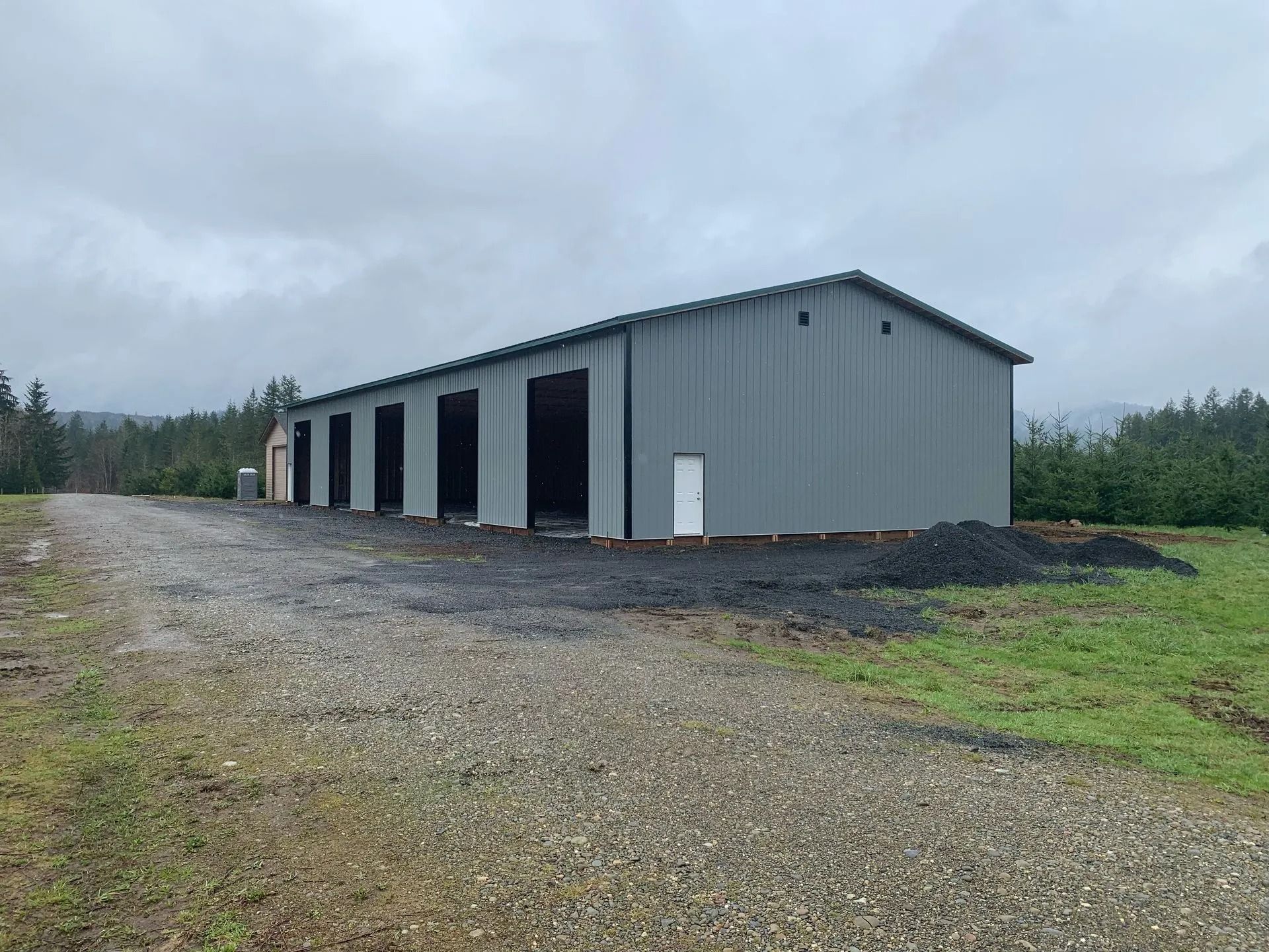 Gray metal building with multiple open bays on a gravel lot under an overcast sky.