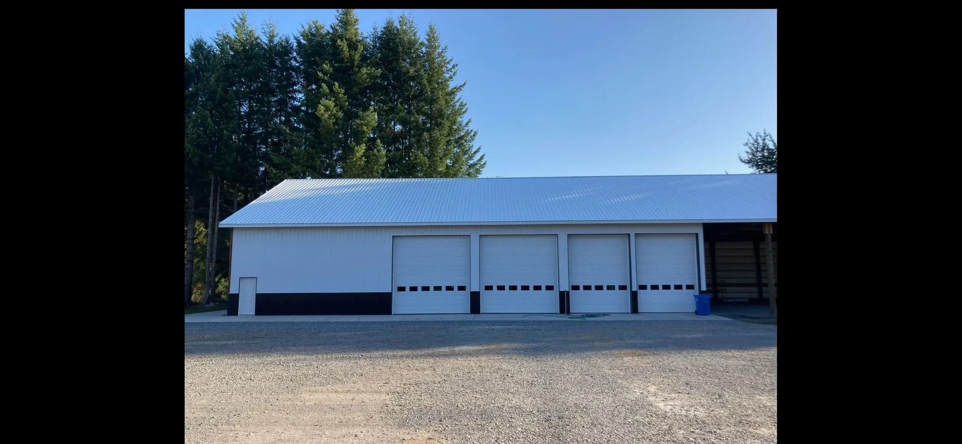 White building with garage doors, snowy roof, gravel ground, trees in background.