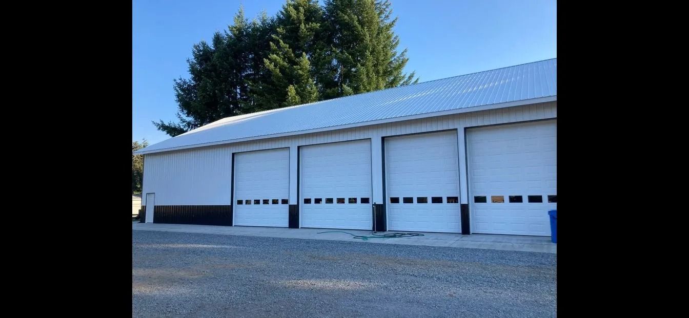 A white building with four closed garage doors under a blue sky, surrounded by trees.
