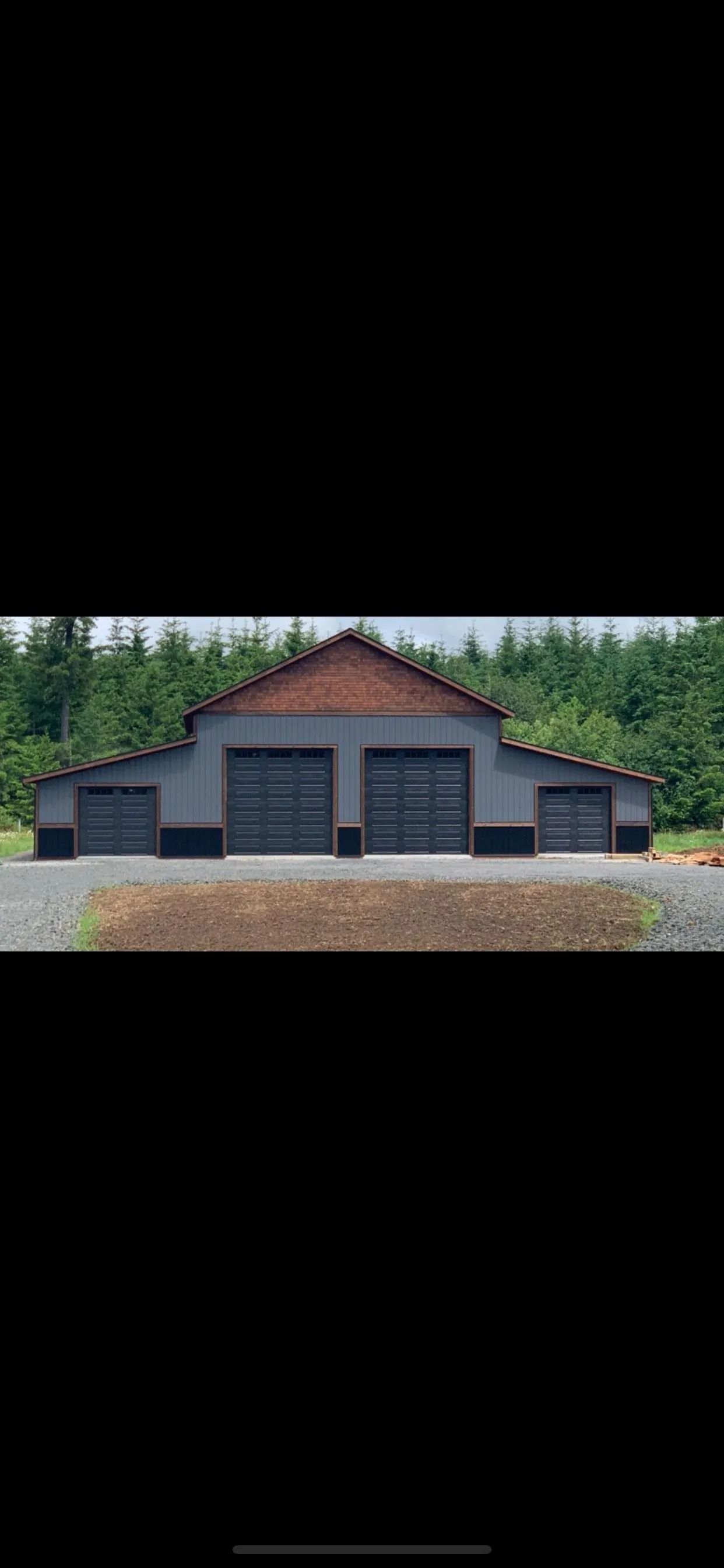Dark gray building with two large and two small garage doors, brown roof, set in front of green trees.