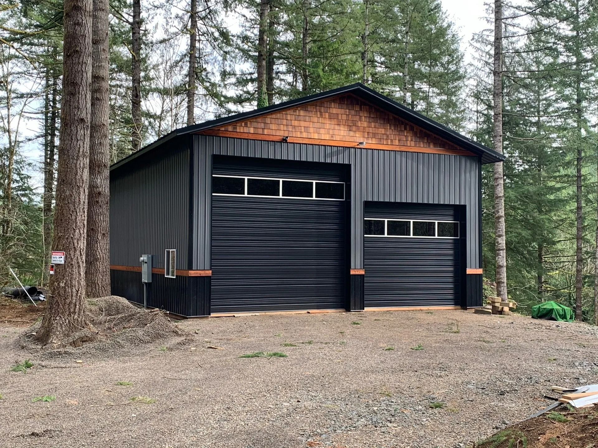 Black metal garage in a forest setting. Two garage doors, wood trim, and a brick accent.