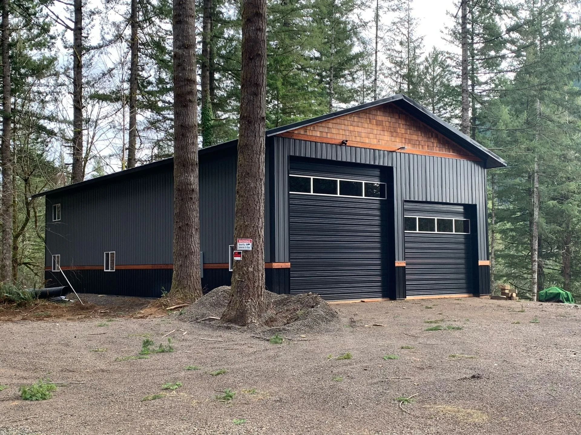 Black metal garage building in wooded area, two garage doors with windows, tan wood trim.
