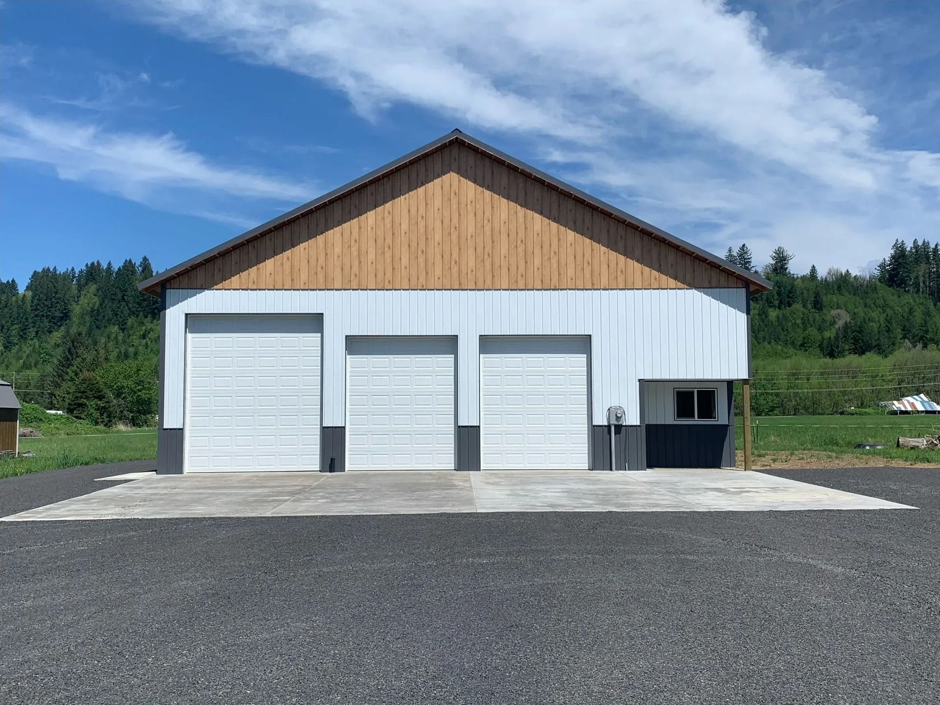 Garage with three white doors, gray trim, and tan wood above, set on gravel. Blue sky background.