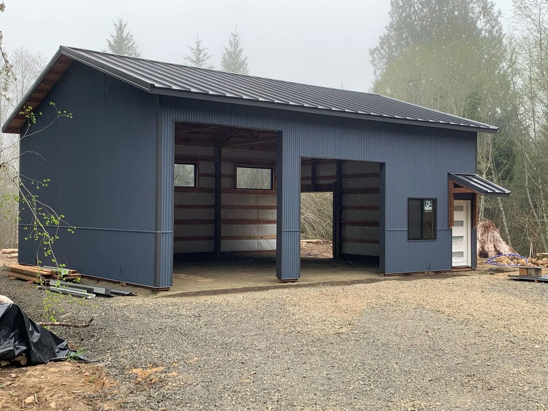 Blue metal building with two open garage doors. Gravel driveway, forested background.