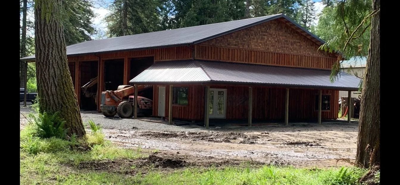 A wooden barn with a dark metal roof sits on a muddy lot with trees in the foreground.