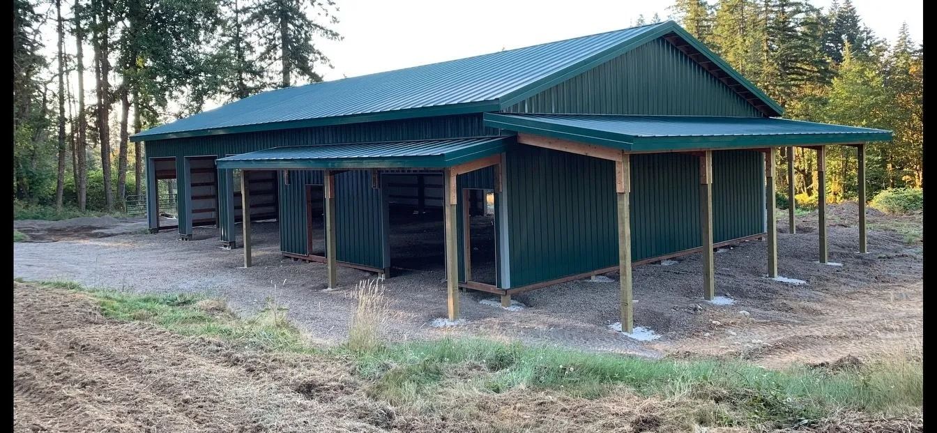 Green metal barn with covered entryway in a rural setting, next to a wooded area.