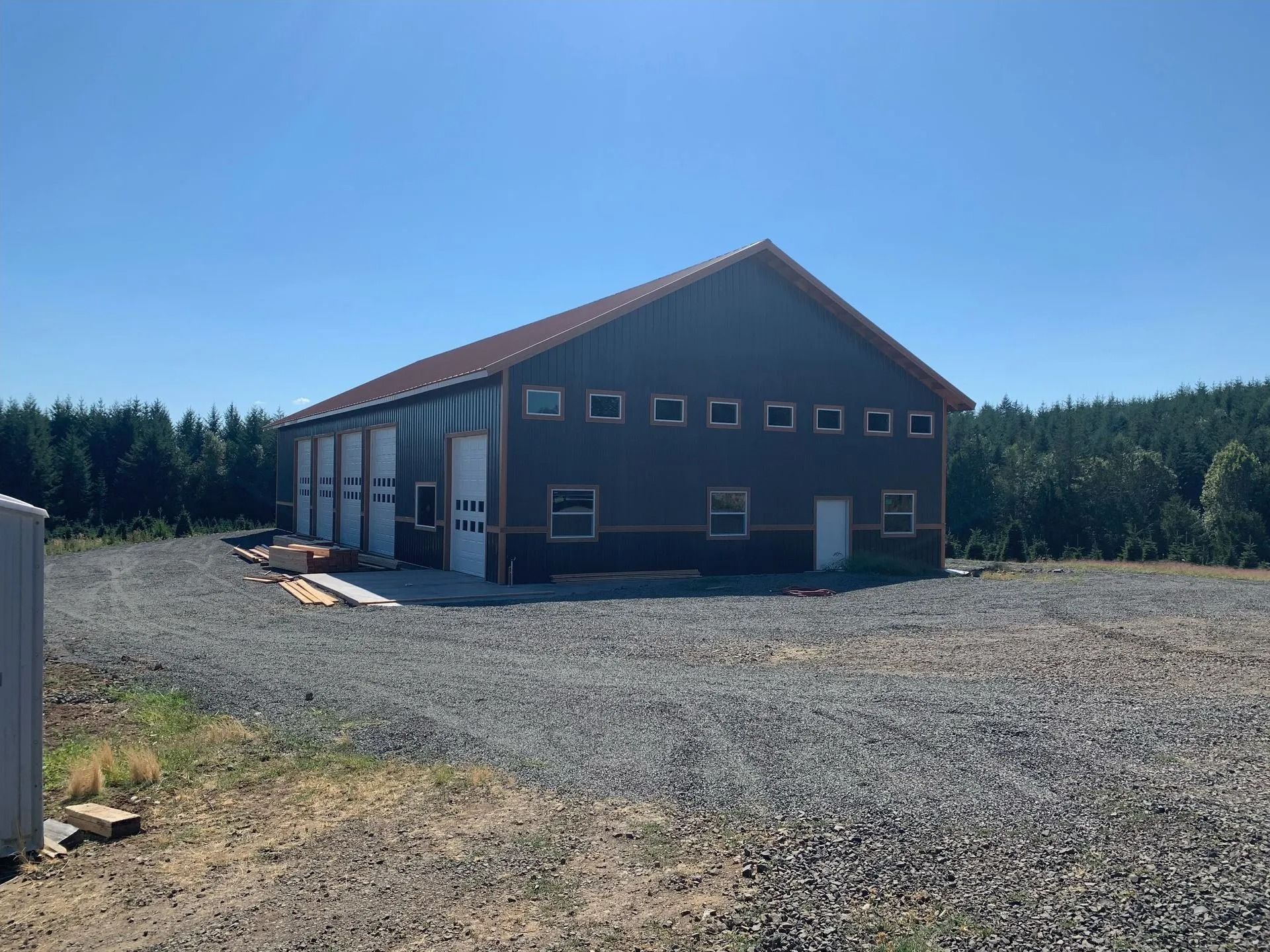 Dark gray metal building with garage doors, small windows, gravel driveway, and trees under a blue sky.